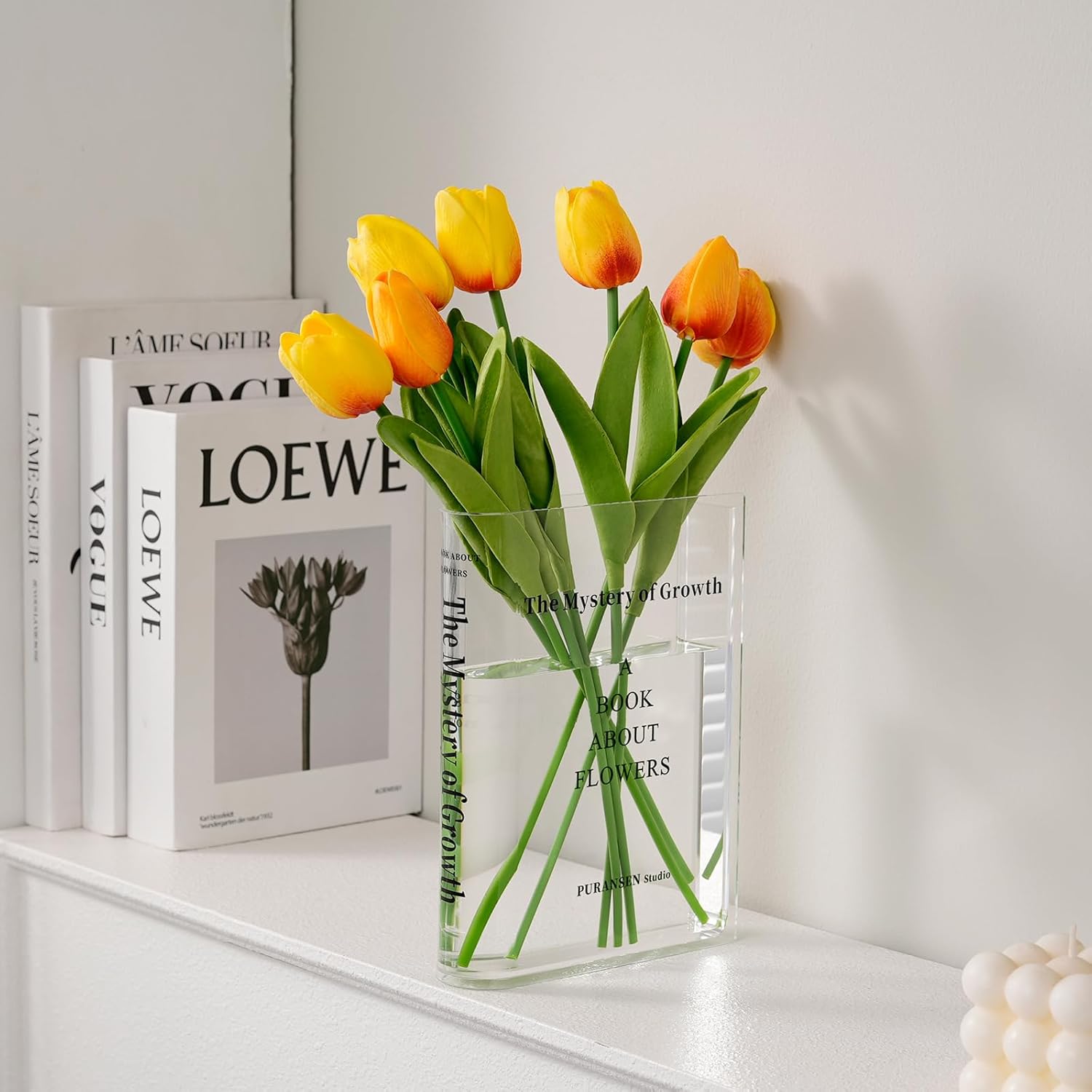 Clear acrylic book-shaped vase with yellow tulips on a white shelf
